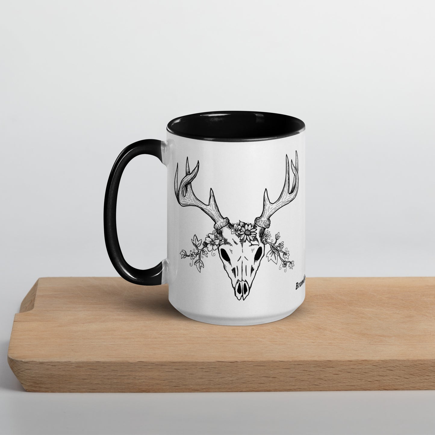 15 ounce ceramic mug with hand illustrated deer skull wreathed in flowers. Double-sided design. Mug has black rim, handle, and inside. Shown sitting on wooden cutting board with handle facing left.