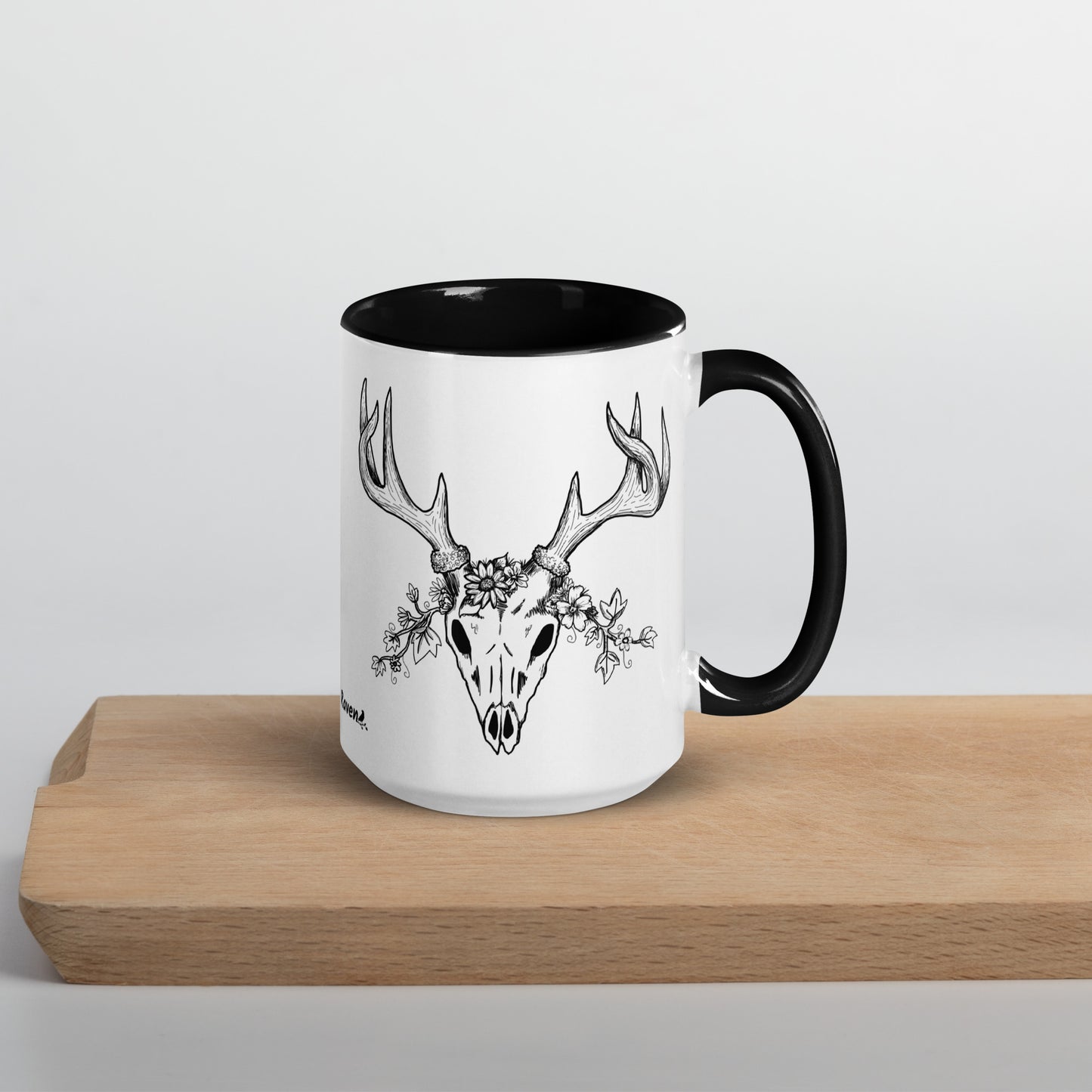 15 ounce ceramic mug with hand illustrated deer skull wreathed in flowers. Double-sided design. Mug has black rim, handle, and inside. Shown sitting on wooden cutting board with handle facing right.