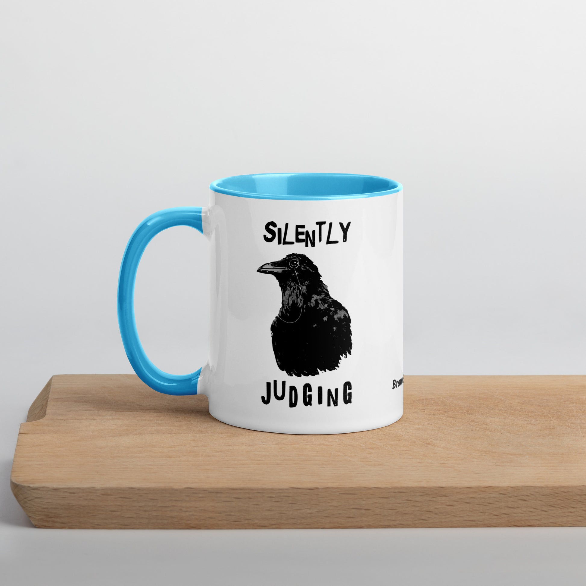 11 ounce ceramic mug with Silently Judging text surrounding a monacle-wearing crow. Double-sided design. Mug has blue rim, handle, and inside. Shown sitting on wooden cutting board.