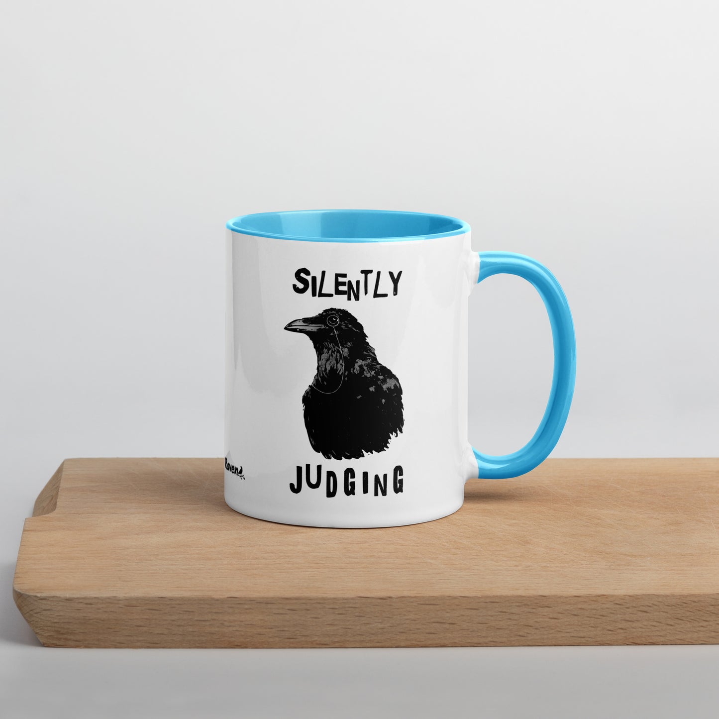 11 ounce ceramic mug with Silently Judging text surrounding a monacle-wearing crow. Double-sided design. Mug has blue rim, handle, and inside. Shown sitting on wooden cutting board with handle facing right.
