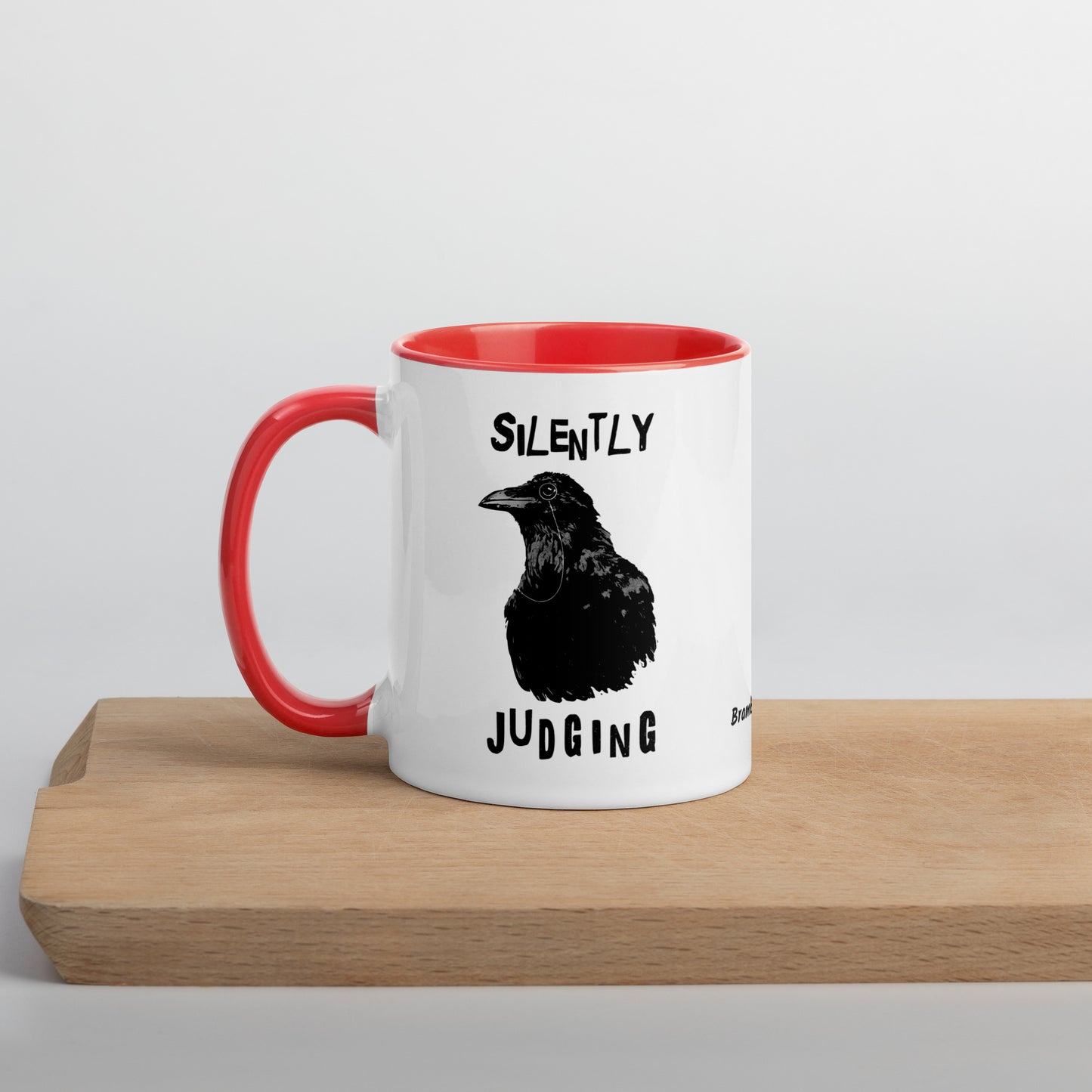11 ounce ceramic mug with Silently Judging text surrounding a monacle-wearing crow. Double-sided design. Mug has red rim, handle, and inside. Shown sitting on wooden cutting board.