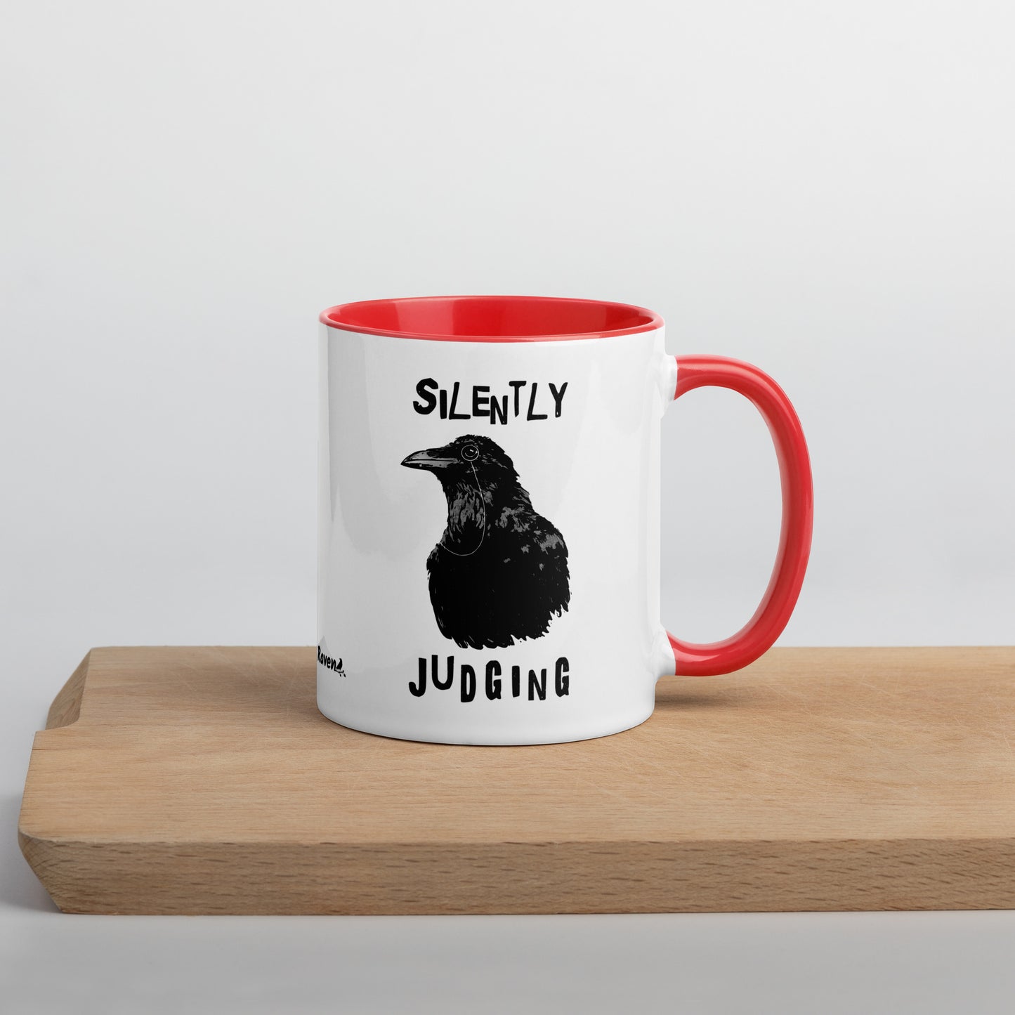 11 ounce ceramic mug with Silently Judging text surrounding a monacle-wearing crow. Double-sided design. Mug has red rim, handle, and inside. Shown sitting on wooden cutting board with handle facing right.