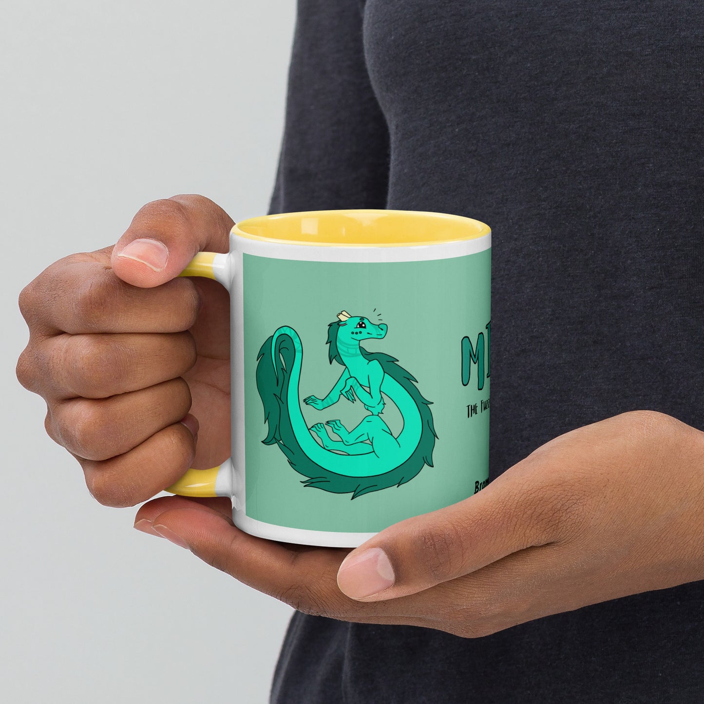 11 ounce white ceramic mug. Yellow inside and handle. Features double-sided image of Minty the Fuzzy Noodle Dragon on a green background. Shown in model's hands.