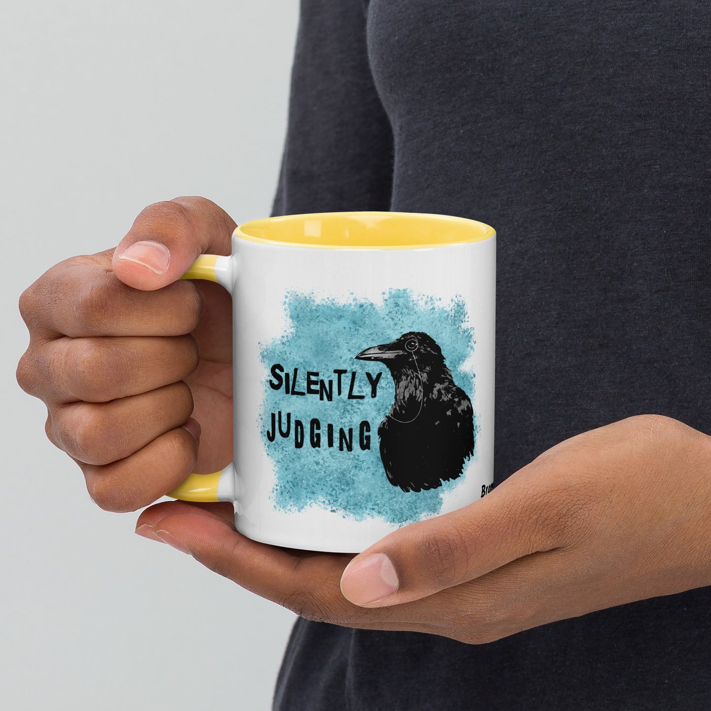11 ounce ceramic mug with Silently Judging text next to a monacle-wearing crow on blue paint splatters. Double-sided design. Mug has yellow rim, handle, and inside. Shown in the hands of a model.