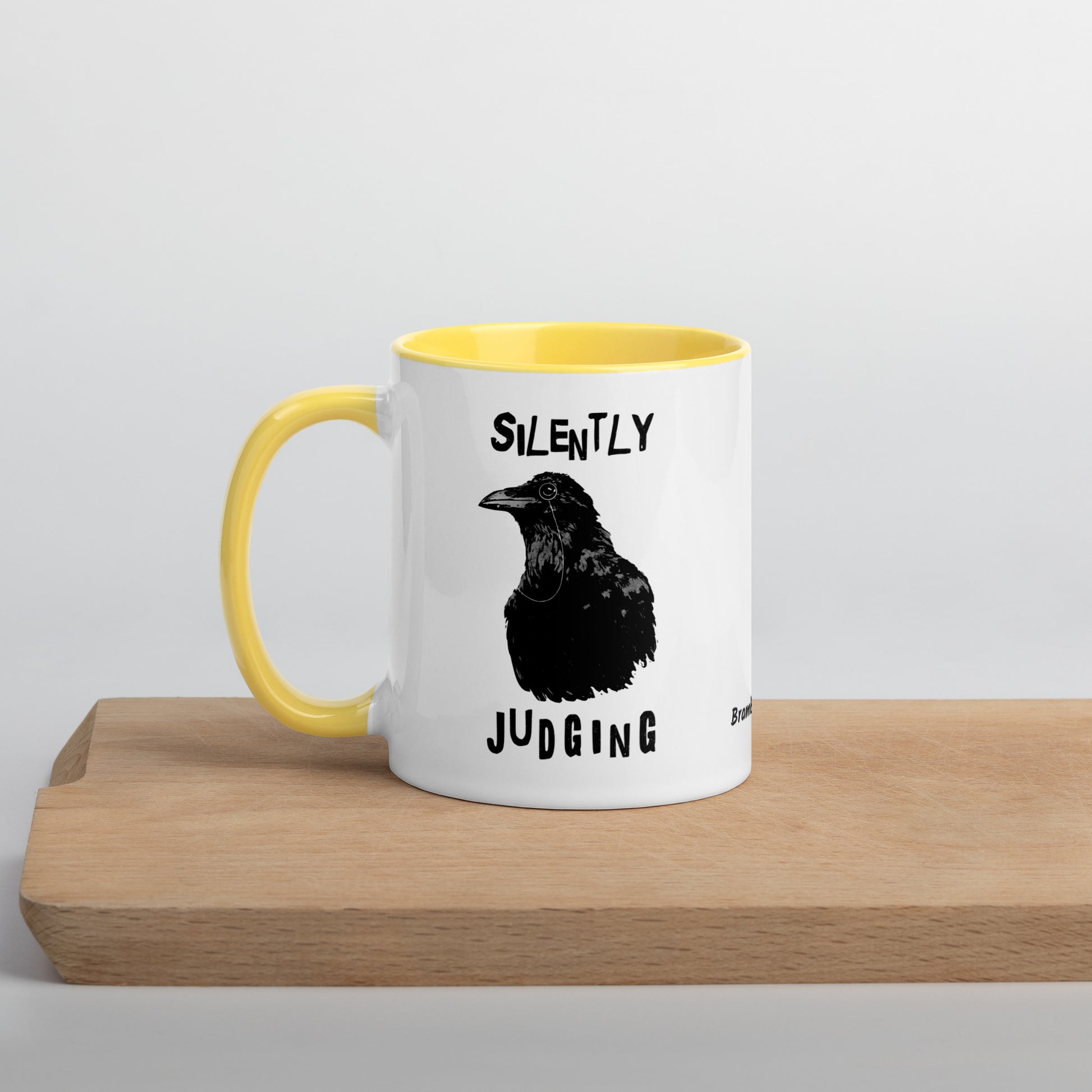11 ounce ceramic mug with Silently Judging text surrounding a monacle-wearing crow. Double-sided design. Mug has yellow rim, handle, and inside. Shown sitting on wooden cutting board.