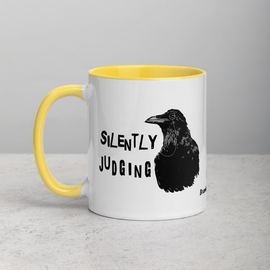 11 ounce ceramic mug with Silently Judging text by a monacle-wearing crow. Double-sided design. Mug has yellow rim, handle, and inside. Shown sitting on a tabletop.