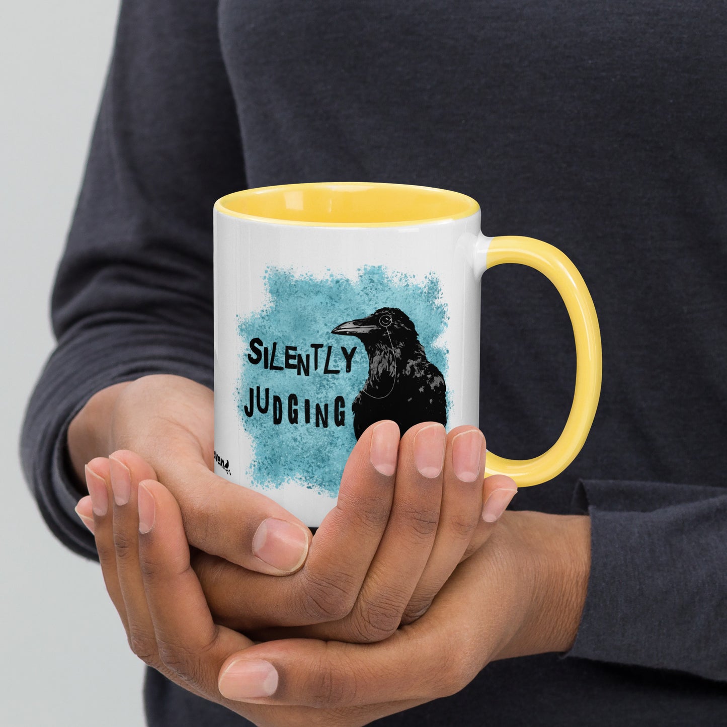 11 ounce ceramic mug with Silently Judging text next to a monacle-wearing crow on blue paint splatters. Double-sided design. Mug has yelllow rim, handle, and inside. Shown in the hands of a model.