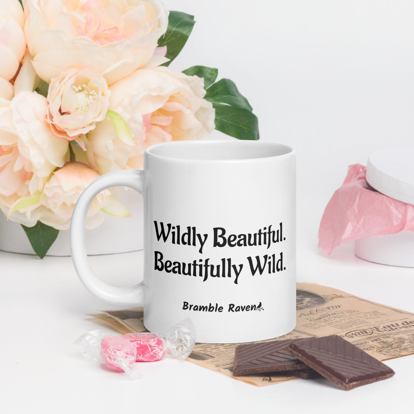 20 ounce white ceramic mug. Features hand-illustrated deer skull wreathed in flowers on one side, and text wildy beautiful, beautifully wild on the other side. Shown by flowers and chocolate.