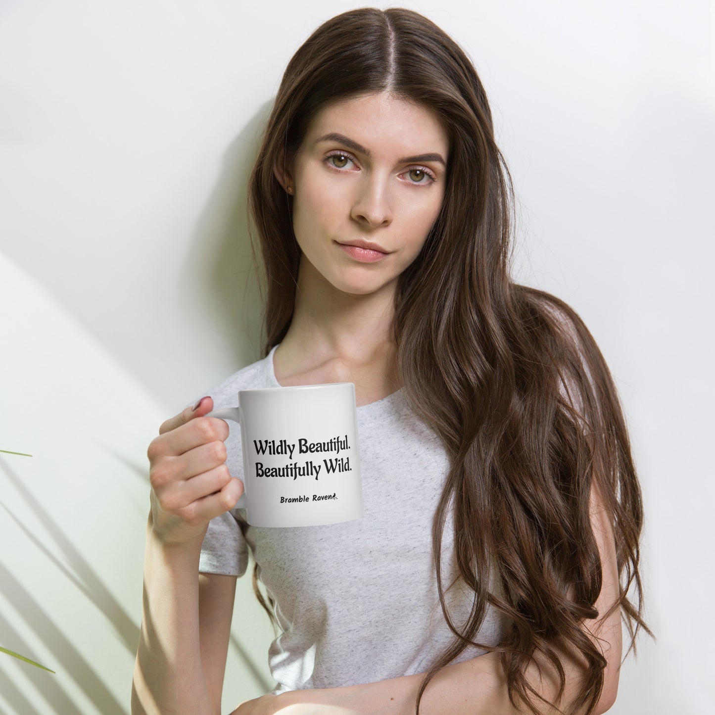 20 ounce white ceramic mug. Features hand-illustrated deer skull wreathed in flowers on one side, and text wildy beautiful, beautifully wild on the other side. Shown in female model's hand.