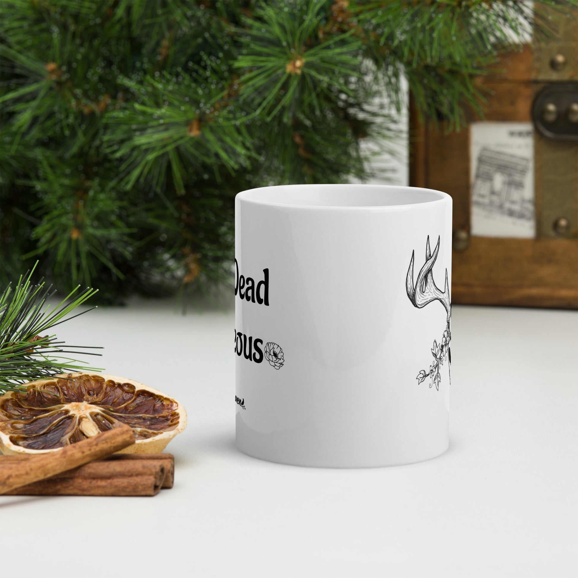 11 ounce white ceramic mug. Features illustrated deer skull wreathed in flowers on one side and Drop dead gorgeous text on the other side. Dishwasher and microwave safe. Front view of mug shown by cinnamon sticks, dried citrus slices, and pine boughs.