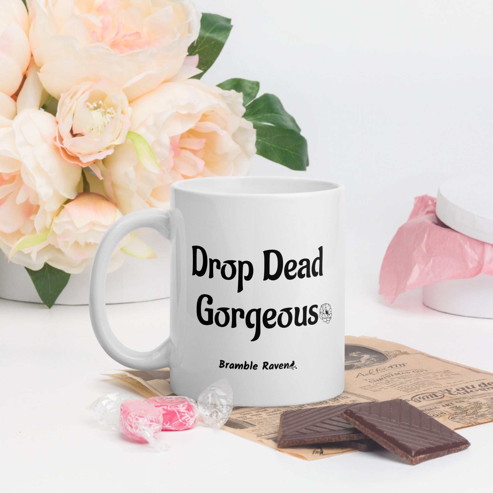 11 ounce white ceramic mug. Features illustrated deer skull wreathed in flowers on one side and Drop dead gorgeous text on the other side. Dishwasher and microwave safe. Shown by flowers and chocolate.