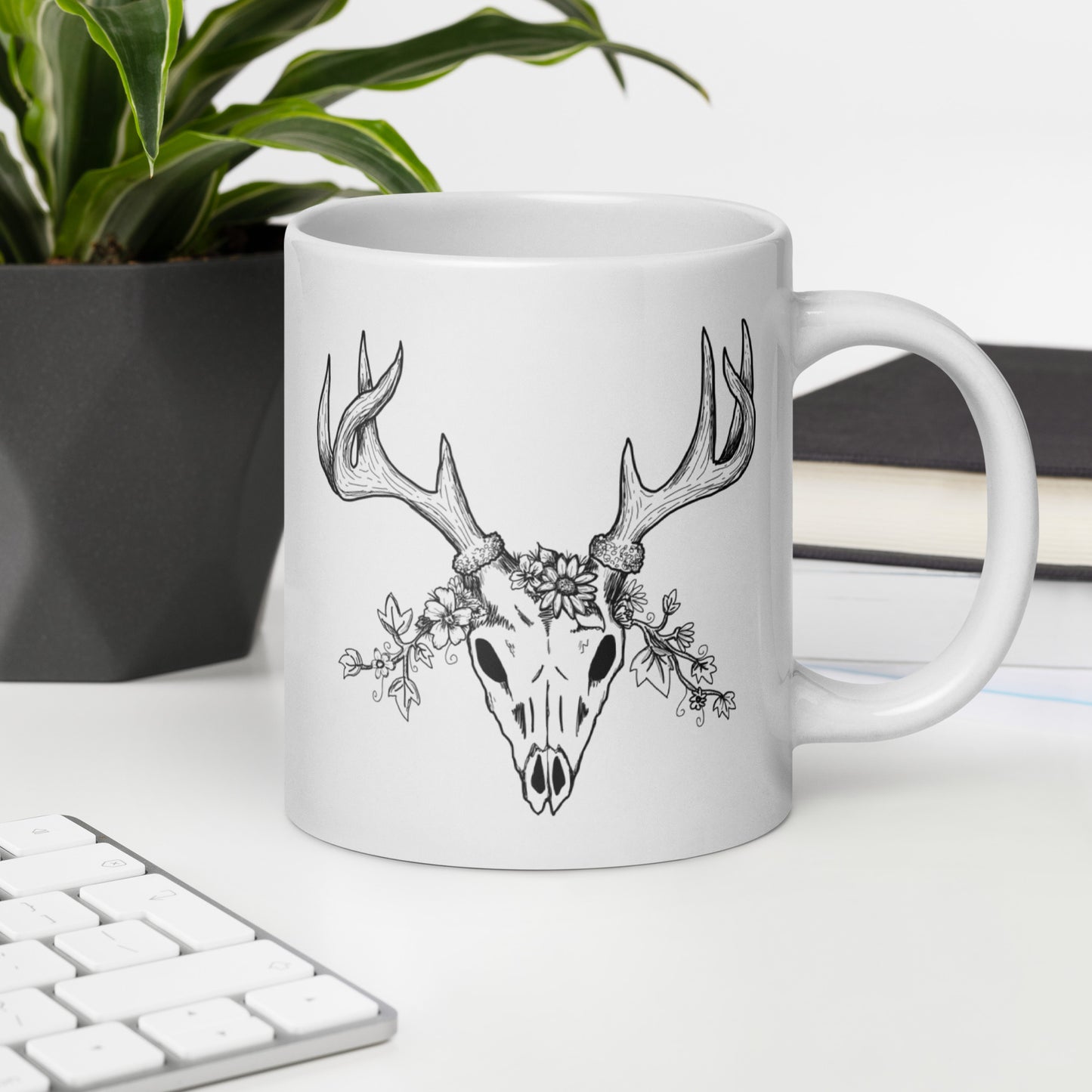 20 ounce white ceramic mug. Features illustrated deer skull wreathed in flowers on one side and Drop dead gorgeous text on the other side. Dishwasher and microwave safe. Shown on desk by keyboard, book, and potted plant.