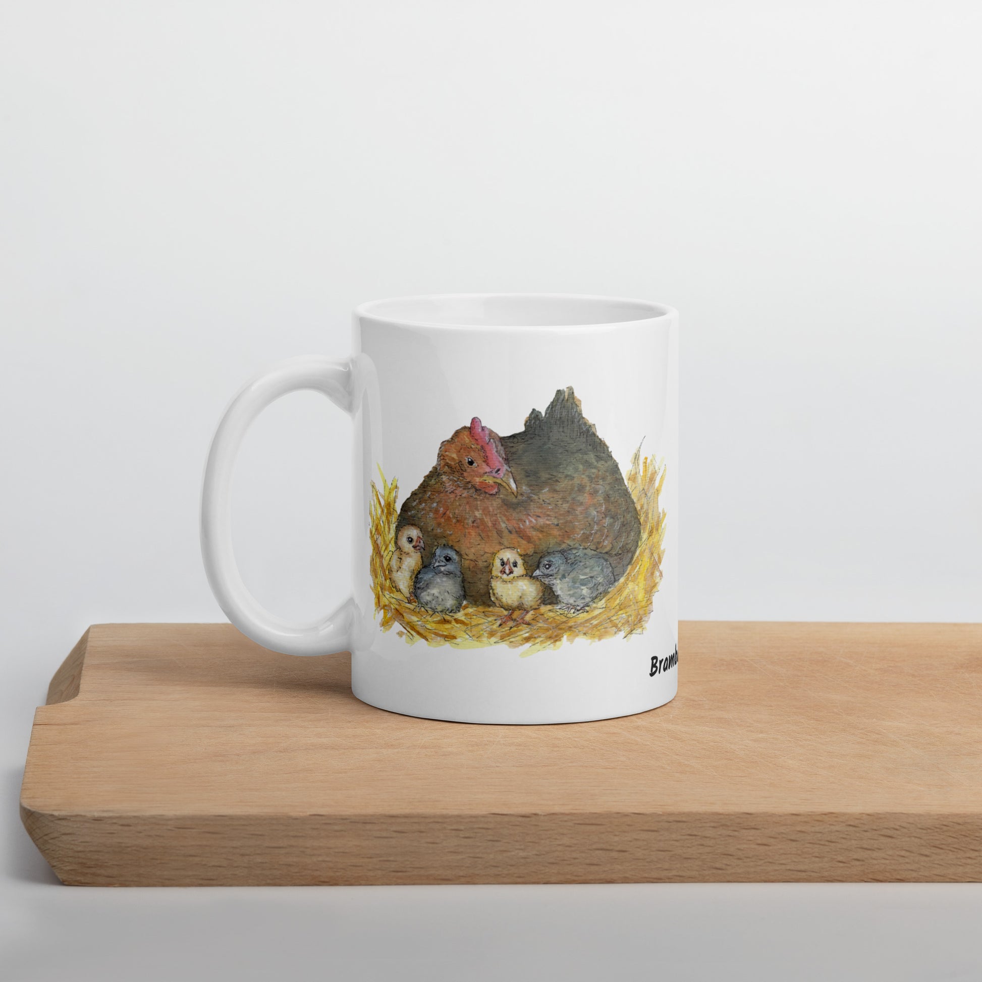11 ounce white ceramic mug. Features double-sided print of a watercolor mother hen and chicks. Dishwasher and microwave safe. Image shows mug sitting on wooden cutting board.