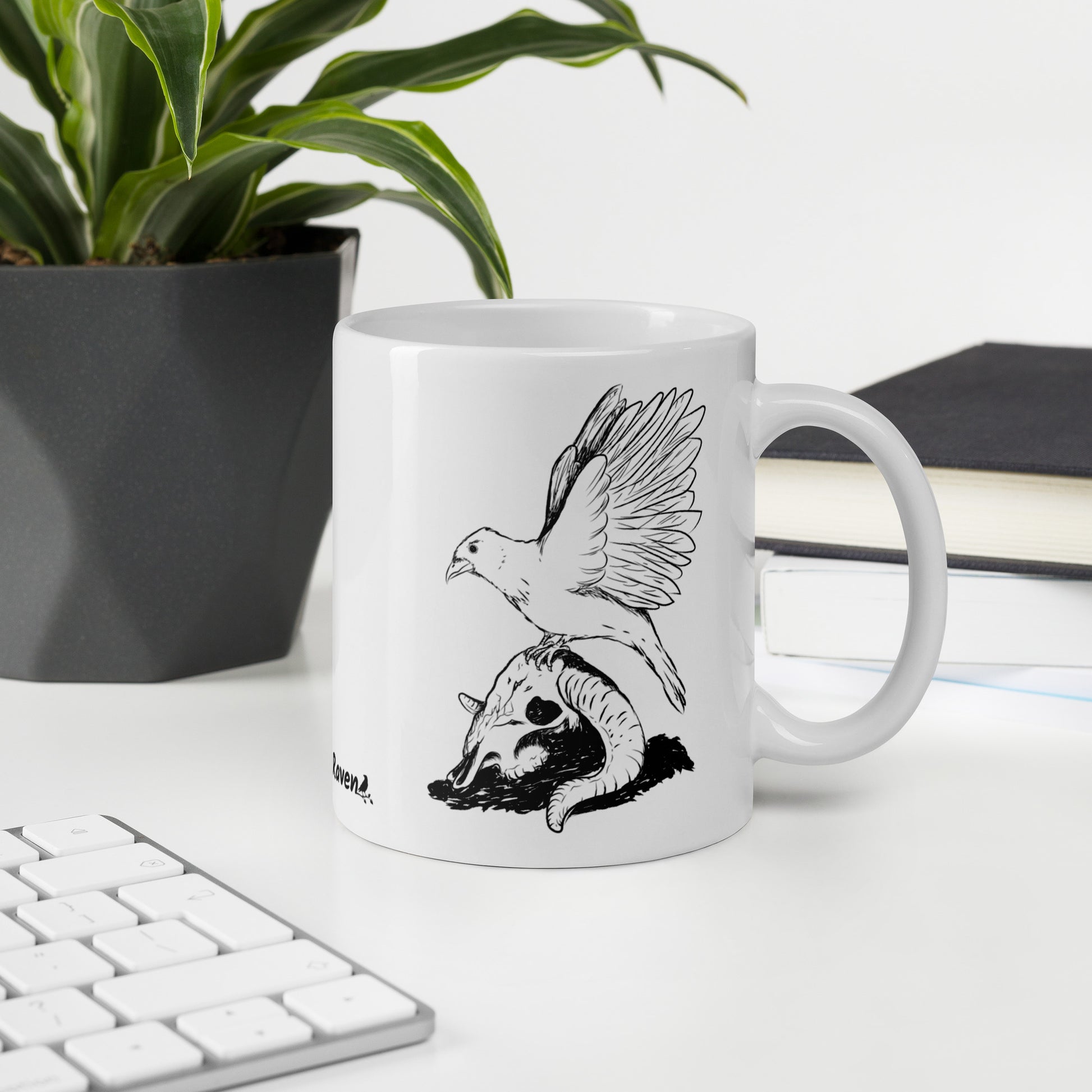 11 ounce white glossy mug featuring double sided print of Reflections, a design with a crow with wings outstretched on a sheep skull. Shown on desktop by plant and keyboard.