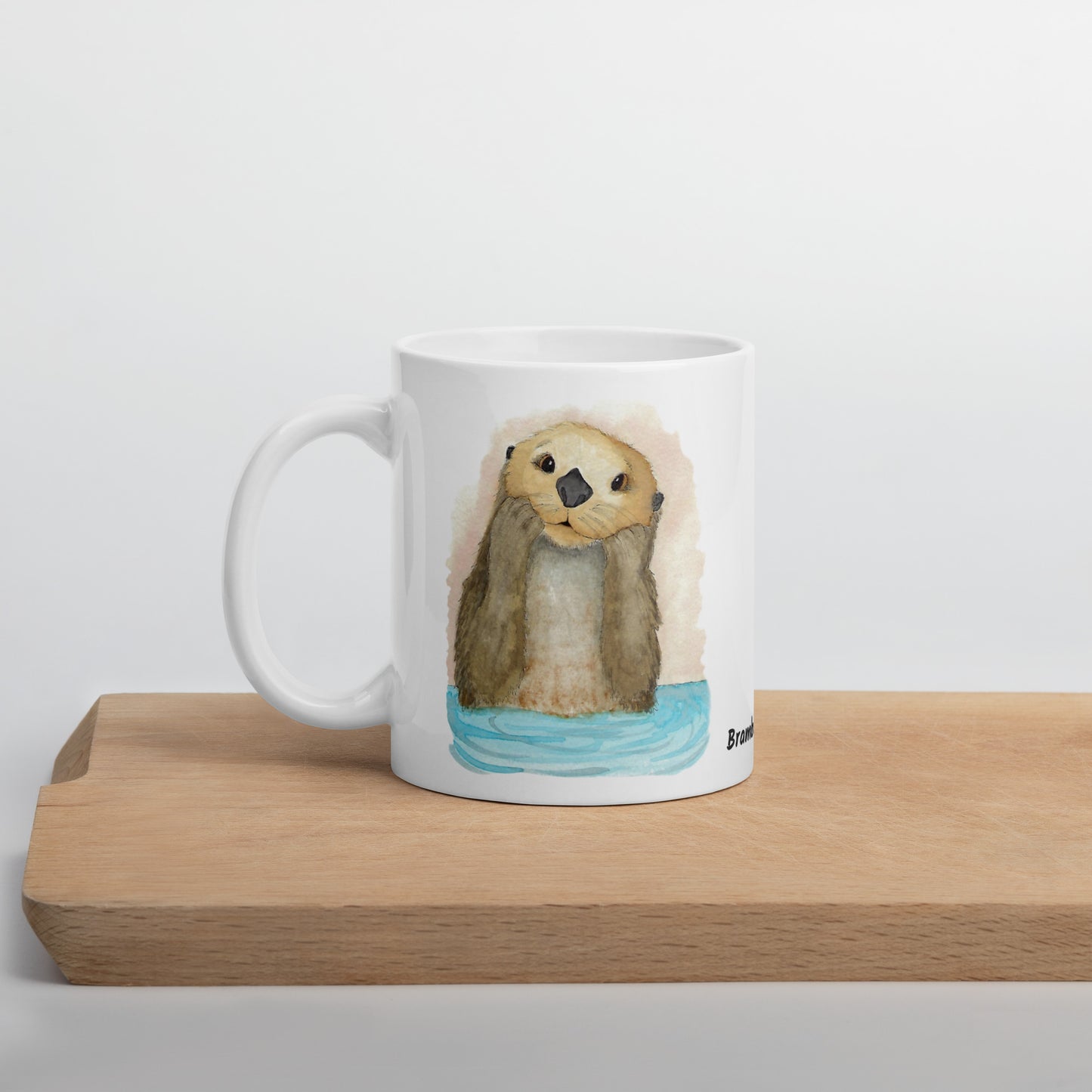 11 ounce white ceramic mug featuring watercolor print of a sea otter on both sides. Dishwasher and microwave safe. Shown on wooden tray.