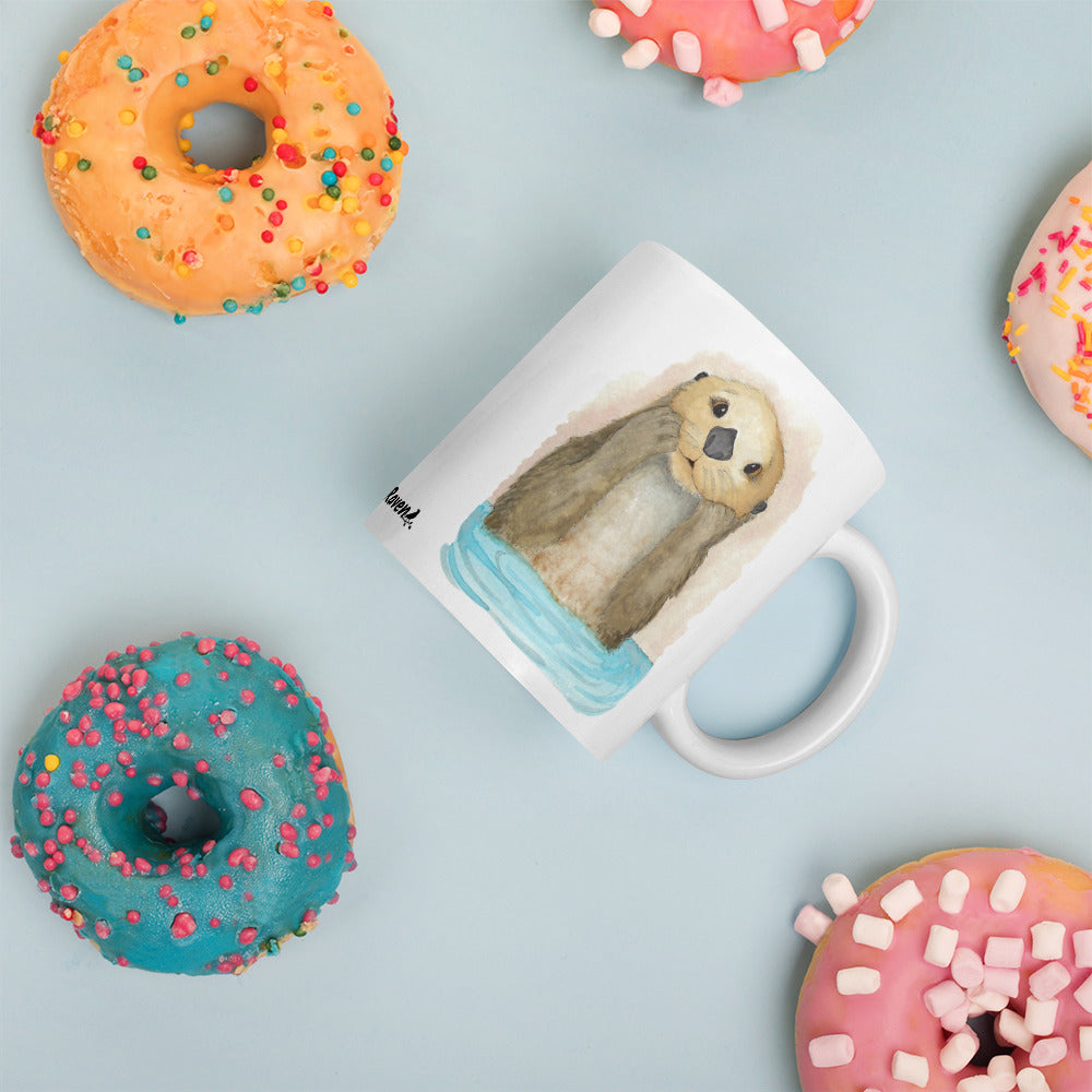 11 ounce white ceramic mug featuring watercolor print of a sea otter on both sides. Dishwasher and microwave safe. Shown laying on tabletop by doughnuts.