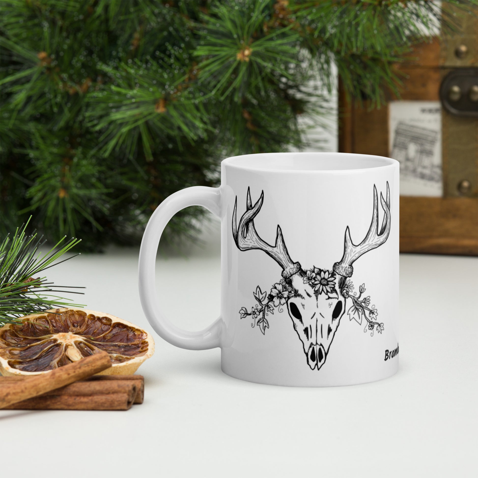 11 oz white ceramic mug. Features a double-sided hand illustrated image of a deer skull wreathed in flowers. Shown on tabletop by pine branches, dried orange slice, and cinnamon sticks.