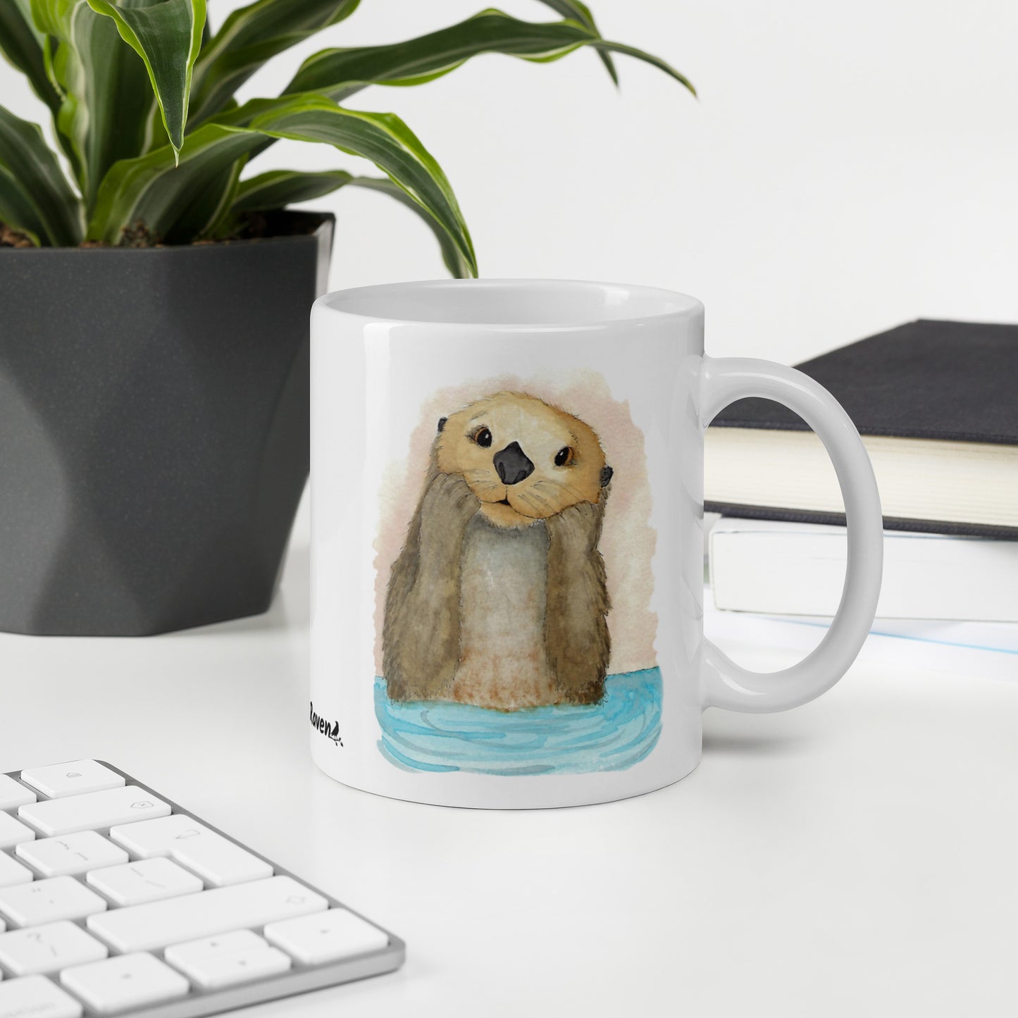 11 ounce white ceramic mug featuring watercolor print of a sea otter on both sides. Dishwasher and microwave safe. Shown on desk by keyboard, book, and potted plant.