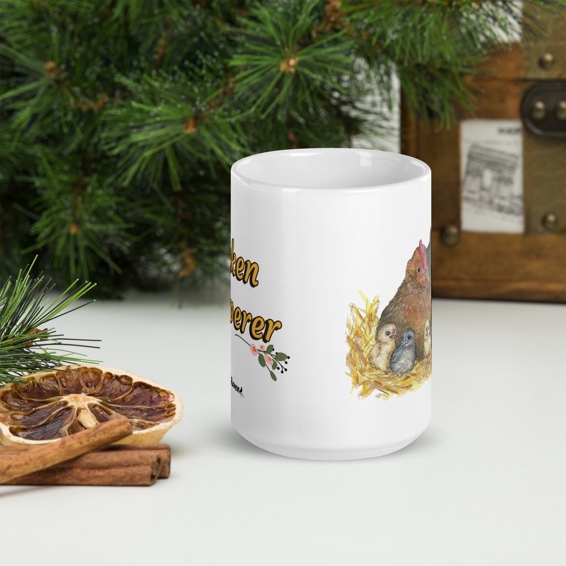 15 ounce white ceramic chicken whisperer mug. Features print of watercolor mother hen and chicks on one side and chicken whisperer text on the other. Dishwasher and microwave safe. Front view shown on tabletop by dried citrus slices and pine boughs.