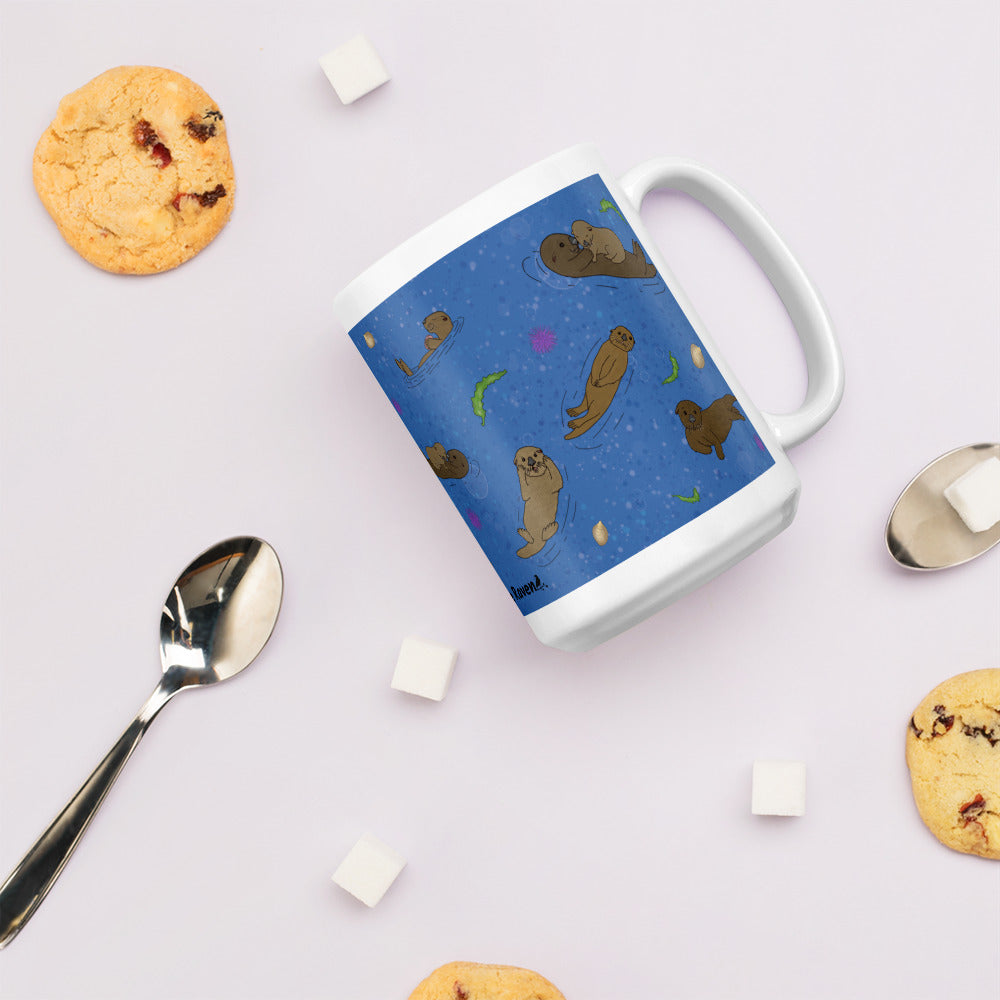 15 ounce white ceramic mug with sea otters on an ocean blue background. Mug is microwave and dishwasher safe. Shown on tabletop by cookies, sugar cubes, and a spoon.