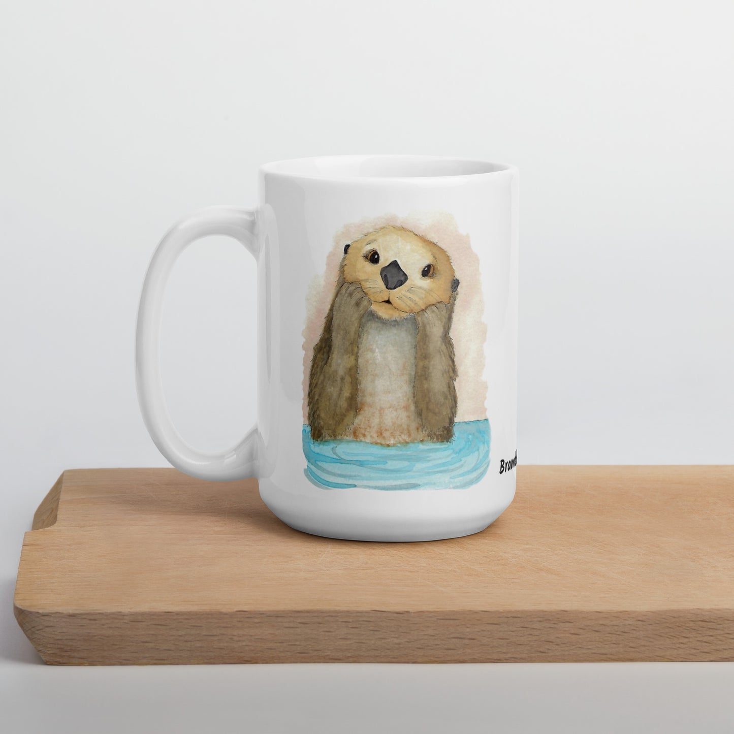 15 ounce white ceramic mug featuring watercolor print of a sea otter on both sides. Dishwasher and microwave safe. Shown on wooden tray.