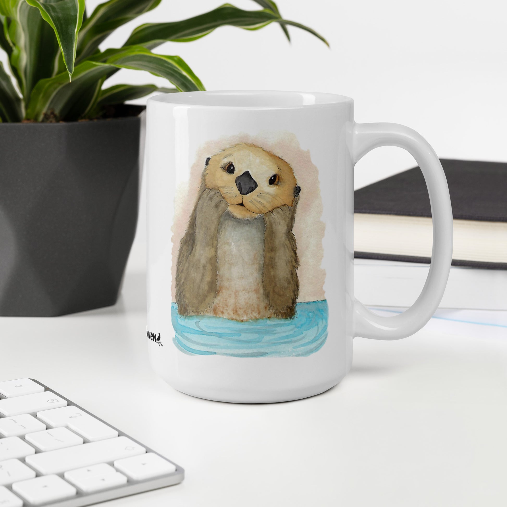 15 ounce white ceramic mug featuring watercolor print of a sea otter on both sides. Dishwasher and microwave safe. Shown on desk by keyboard, book, and potted plant.