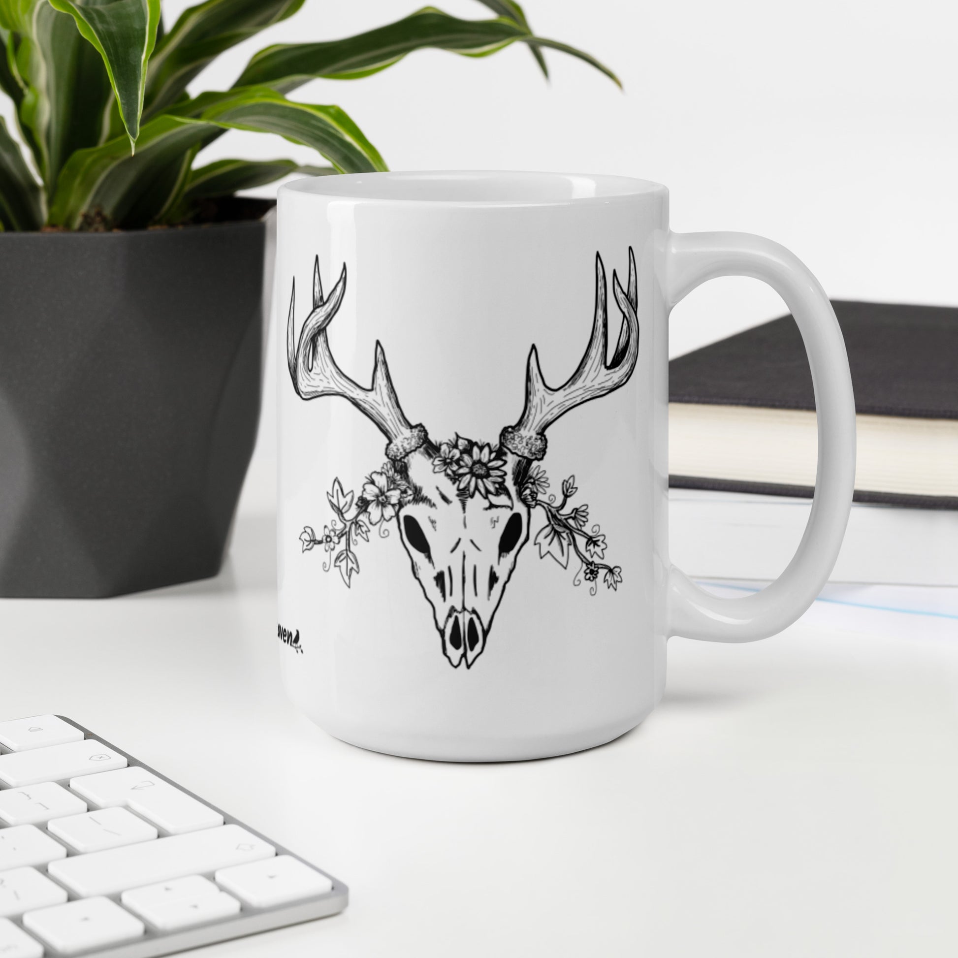 15 oz white ceramic mug. Features a double-sided hand illustrated image of a deer skull wreathed in flowers. Shown on desktop by keyboard, potted plant, and books.