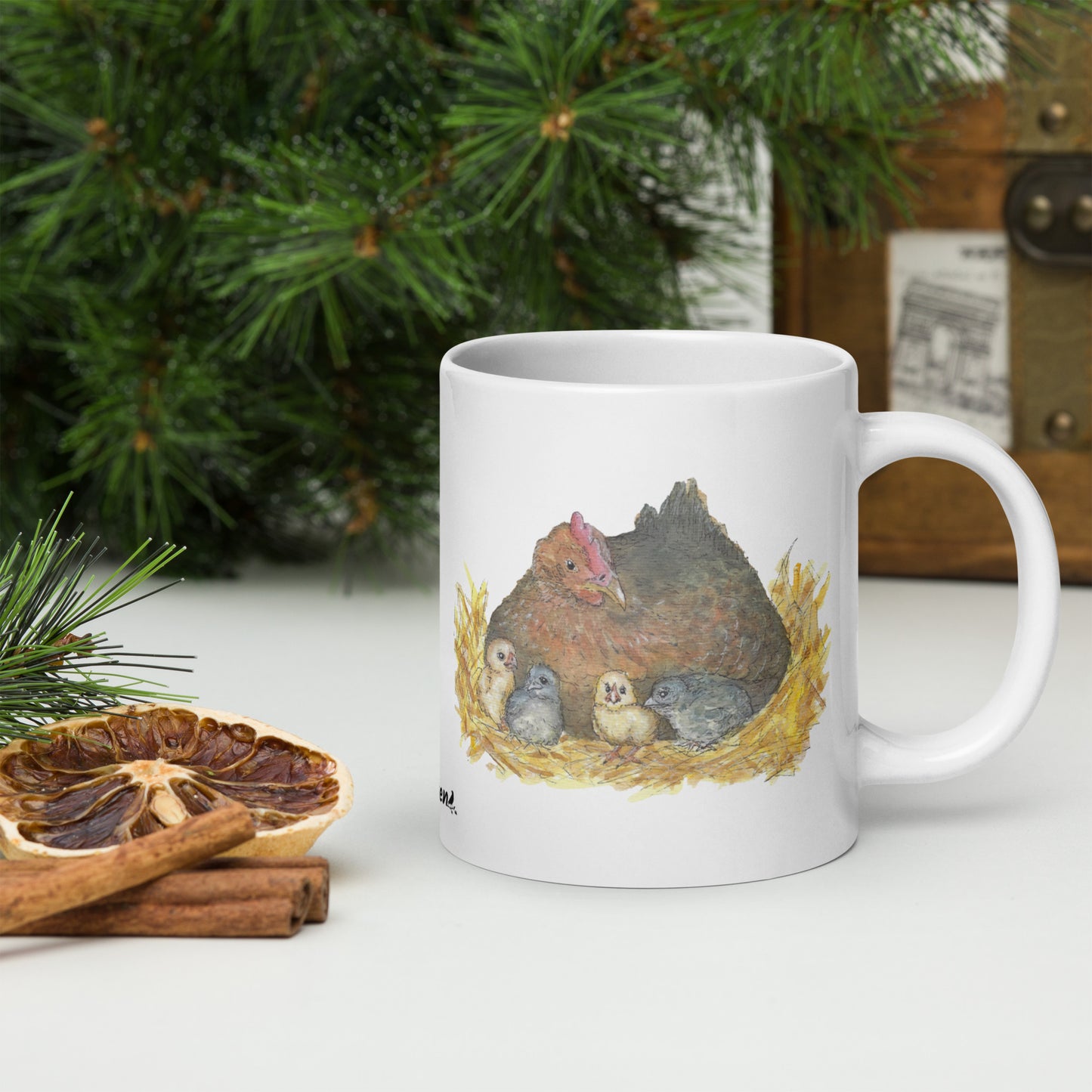 20 ounce white ceramic mug. Features double-sided print of a watercolor mother hen and chicks. Dishwasher and microwave safe. Shown sitting by dried citrus slices and pine boughs.