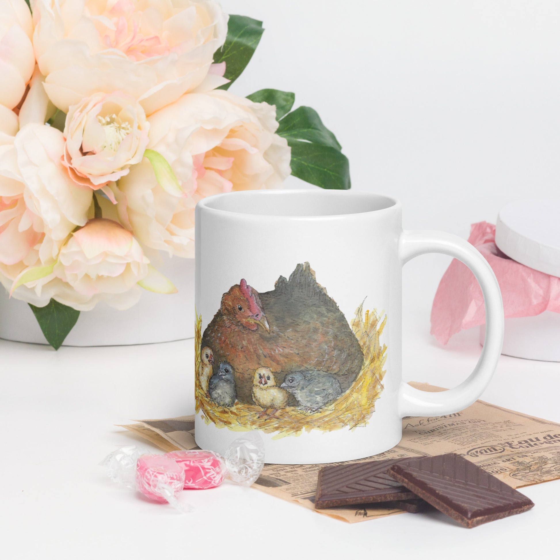 20 ounce white ceramic chicken whisperer mug. Features print of watercolor mother hen and chicks on one side and chicken whisperer text on the other. Dishwasher and microwave safe. Shown on table by chocolate and flowers with handle facing right.
