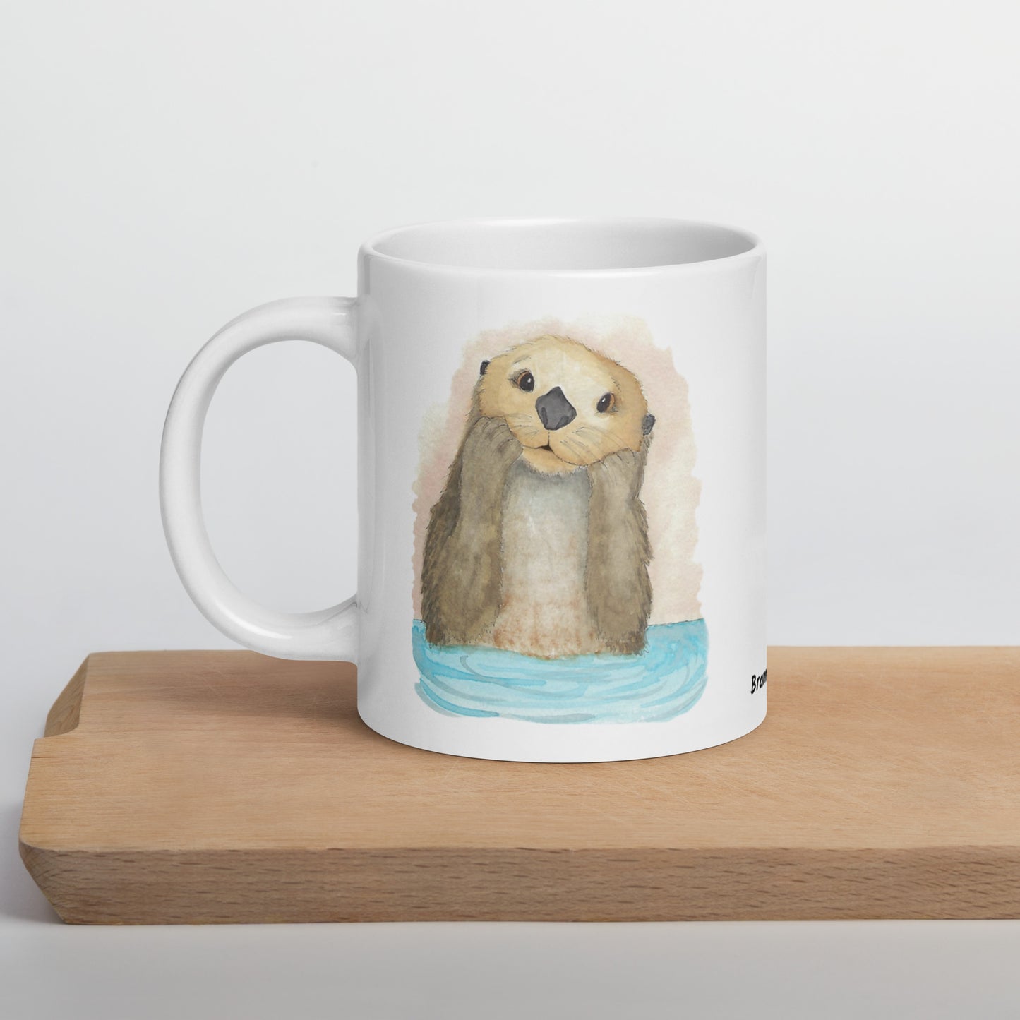 20 ounce white ceramic mug featuring watercolor print of a sea otter on both sides. Dishwasher and microwave safe. Shown on wooden tray.