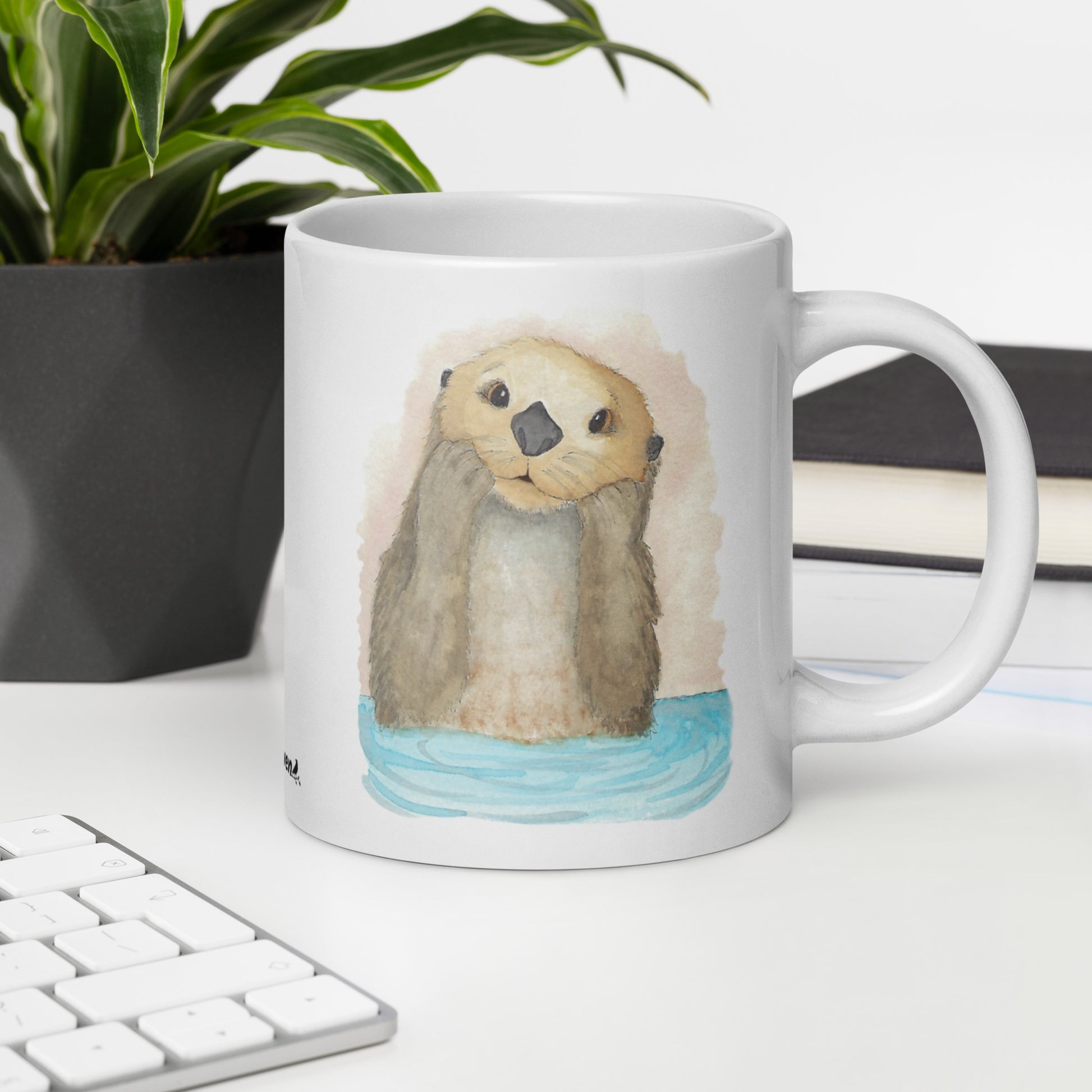 20 ounce white ceramic mug featuring watercolor print of a sea otter on both sides. Dishwasher and microwave safe. Shown on desk by keyboard, book, and potted plant.