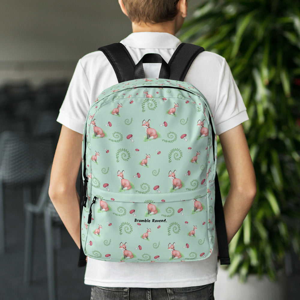 Forest fox backpack. Has watercolor foxes, mushrooms and ferns on a light green water-resistant polyester fabric. Includes an inner pocket that can hold a 15 inch laptop. Has a hidden pocket with zipper on the back, and ergonomic straps. Shown on the back of a boy.
