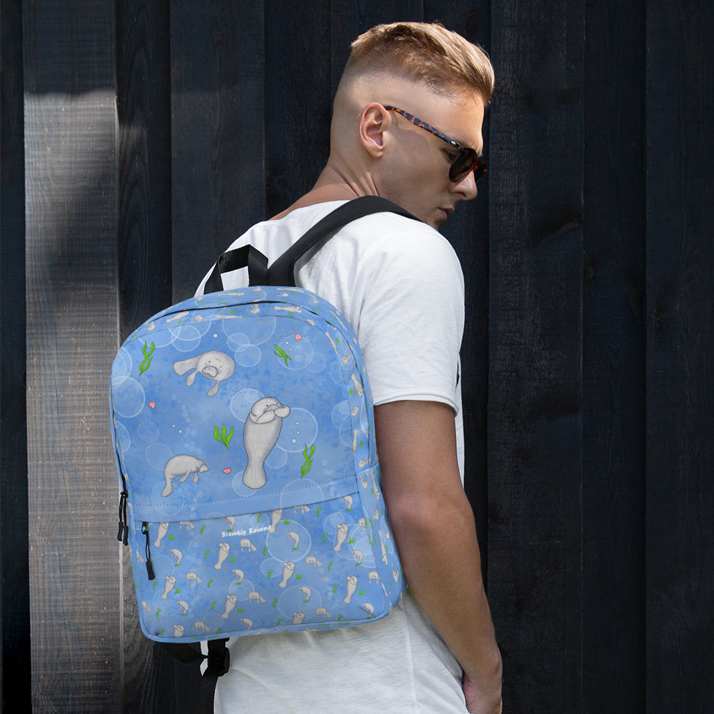 Medium-sized backpack featuring manatees, seashells, seaweed and bubbles on a blue background. Made with water resistant polyester, ergonomic straps, and comfort mesh back. Has a compartment that can hold a 15 inch laptop. Has a hidden zipper pocket on the back. Shown on a male model.