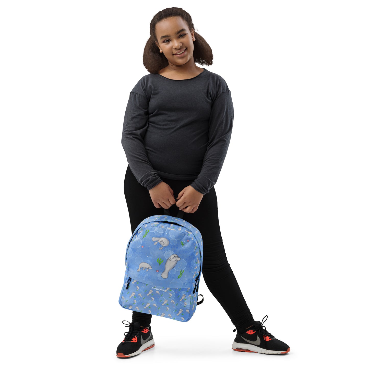 Medium-sized backpack featuring manatees, seashells, seaweed and bubbles on a blue background. Made with water resistant polyester, ergonomic straps, and comfort mesh back. Has a compartment that can hold a 15 inch laptop. Has a hidden zipper pocket on the back. Shown being held by a girl.