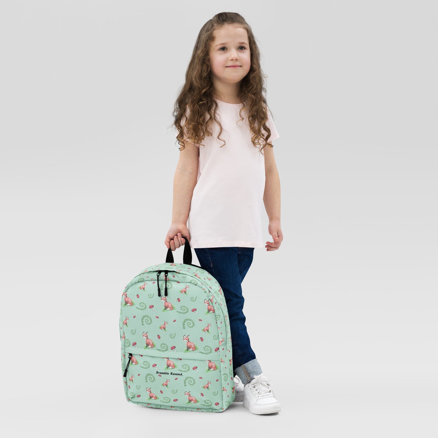 Forest fox backpack. Has watercolor foxes, mushrooms and ferns on a light green water-resistant polyester fabric. Includes an inner pocket that can hold a 15 inch laptop. Has a hidden pocket with zipper on the back, and ergonomic straps. Shown being held by a girl.