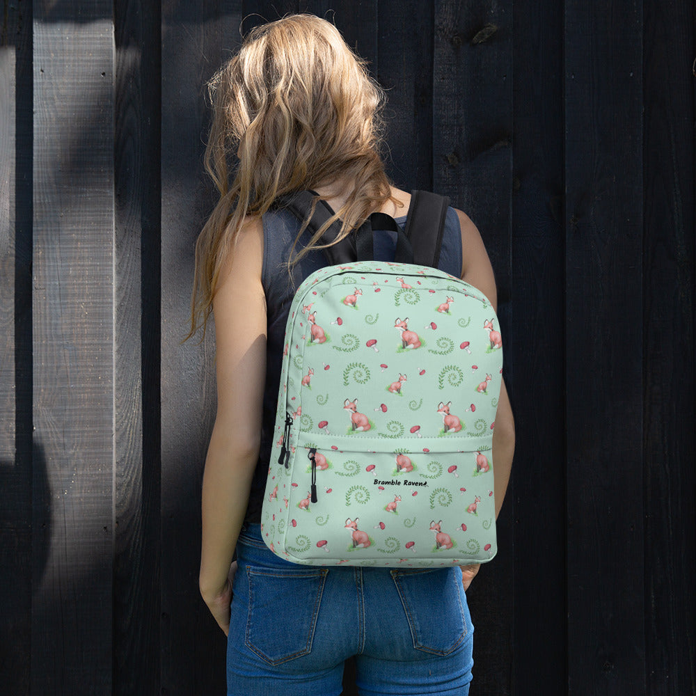Forest fox backpack. Has watercolor foxes, mushrooms and ferns on a light green water-resistant polyester fabric. Includes an inner pocket that can hold a 15 inch laptop. Has a hidden pocket with zipper on the back, and ergonomic straps. Shown on the back of a female model.