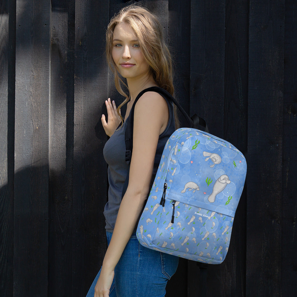 Medium-sized backpack featuring manatees, seashells, seaweed and bubbles on a blue background. Made with water resistant polyester, ergonomic straps, and comfort mesh back. Has a compartment that can hold a 15 inch laptop. Has a hidden zipper pocket on the back. Shown on a female model.