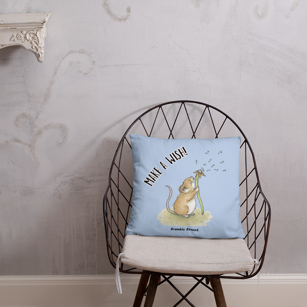 Original Dandelion Wish design of cute watercolor mouse blowing dandelion seeds on a light blue background. Double-sided image on 18 by 18 inch pillow. Back image has make a wish phrase. Shown on  wicker chair.
