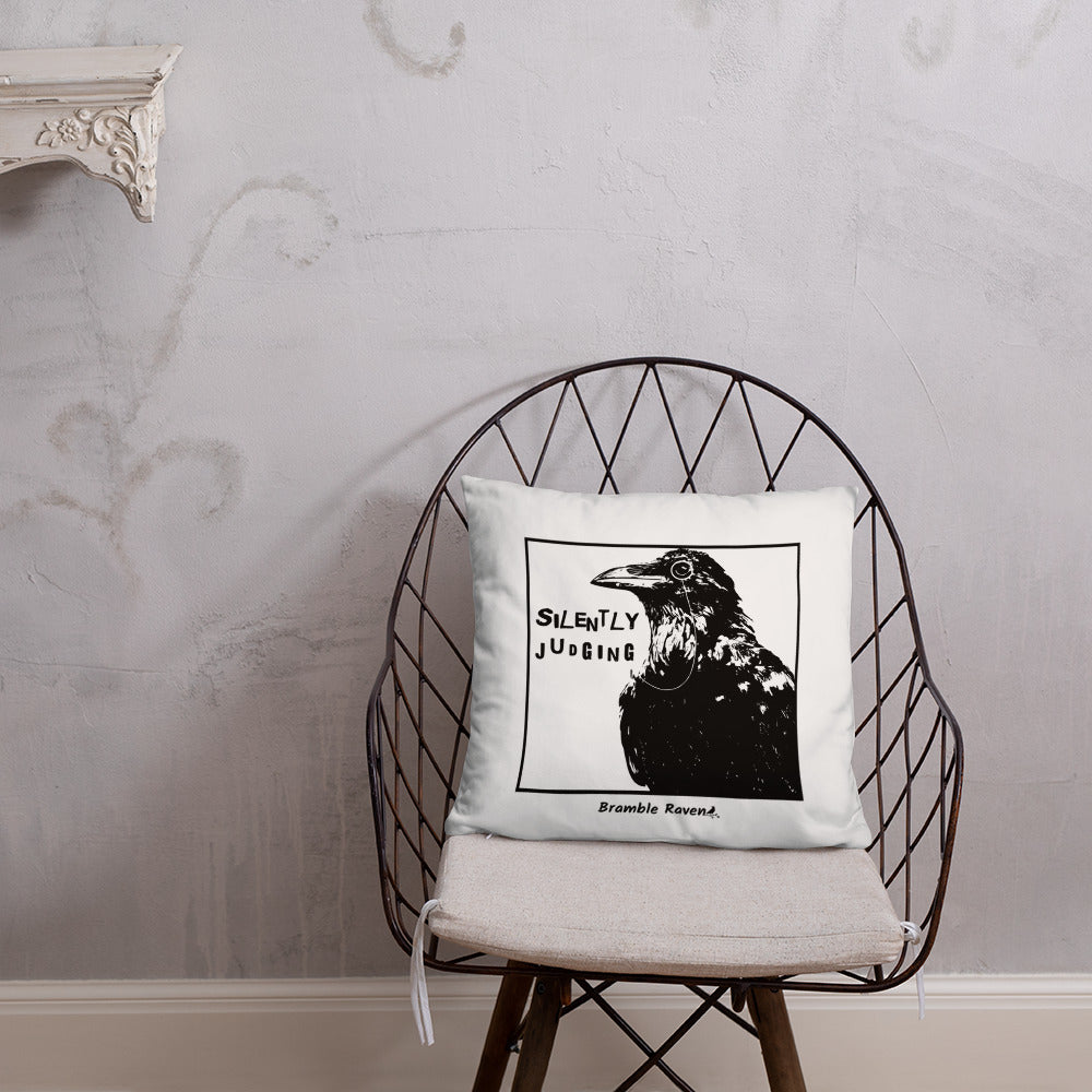 18 by 18 inch accent pillow with silently judging text by black crow wearing a monocle in a square on a white background.  Design has blue paint splatters on reverse side of pillow. Shown on wicker chair.