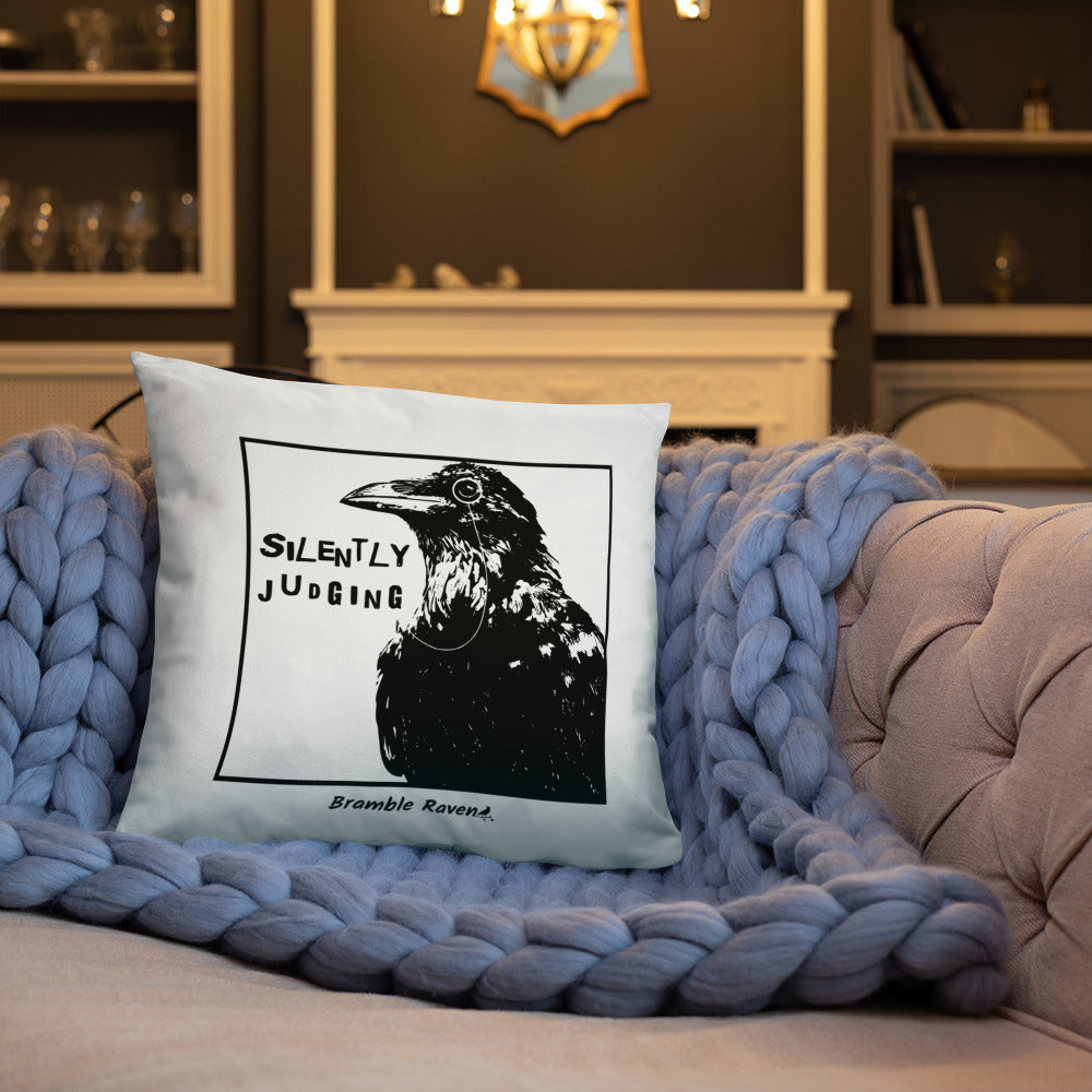 18 by 18 inch accent pillow with silently judging text by black crow wearing a monocle in a square on a white background.  Design reverse side of pillow has blue paint splatters. Shown on blue knitted blanket on tan sofa.