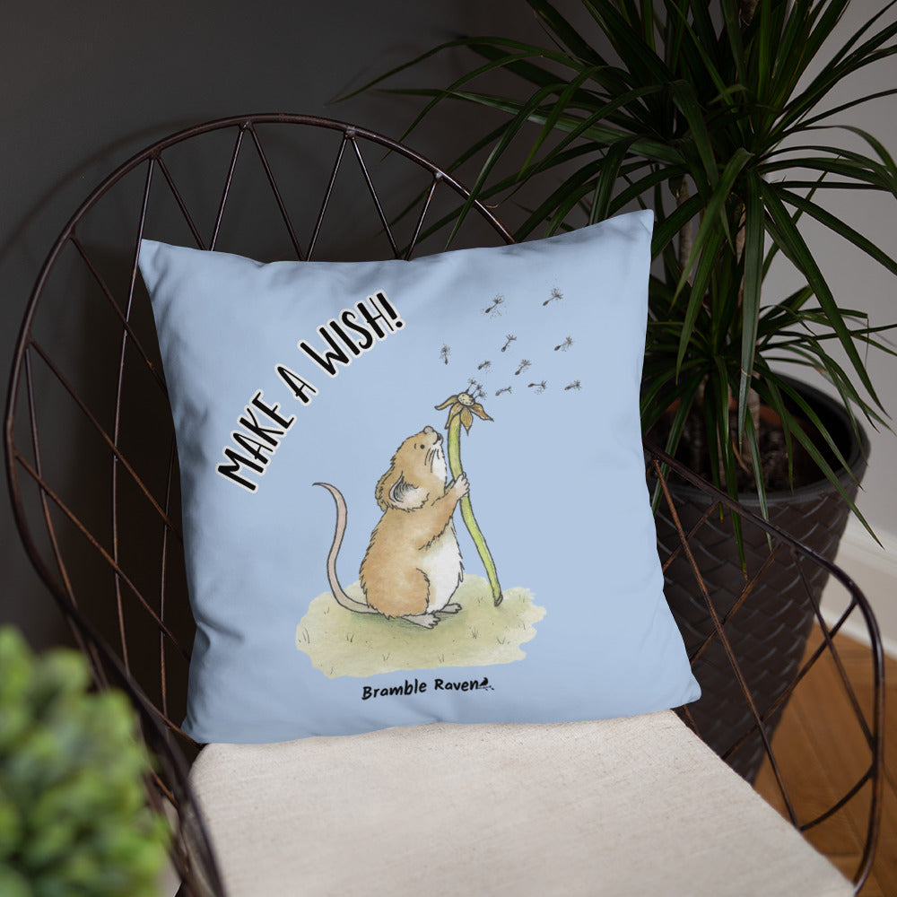 Original Dandelion Wish design of cute watercolor mouse blowing dandelion seeds on a light blue background. Double-sided image on 18 by 18 inch pillow. Back image has make a wish phrase. Shown on wicker chair with potted plant.