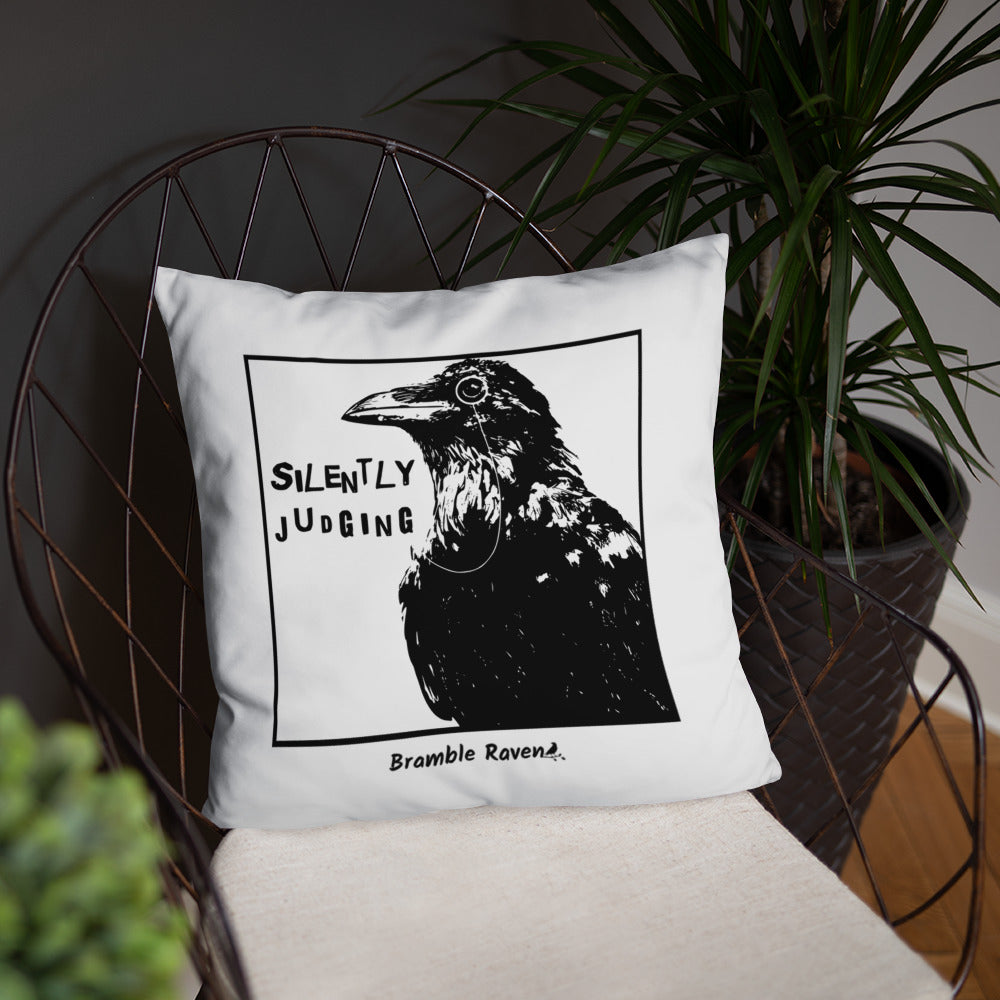 18 by 18 inch accent pillow with silently judging text by black crow wearing a monocle in a square on a white background.  Design has blue paint splatters on reverse side of pillow. Shown on wicker chair.