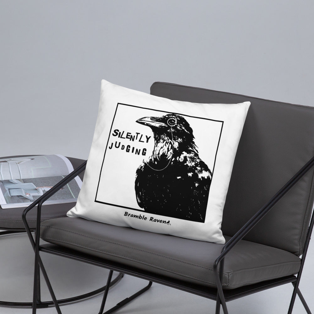 18 by 18 inch accent pillow with silently judging text by black crow wearing a monocle in a square on a white background.  Design on reverse side of pillow has blue paint splatters. Shown on gray chair.