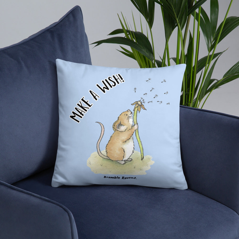Original Dandelion Wish design of cute watercolor mouse blowing dandelion seeds on a light blue background. Double-sided image on 18 by 18 inch pillow. Back image has make a wish phrase. Shown on blue couch.