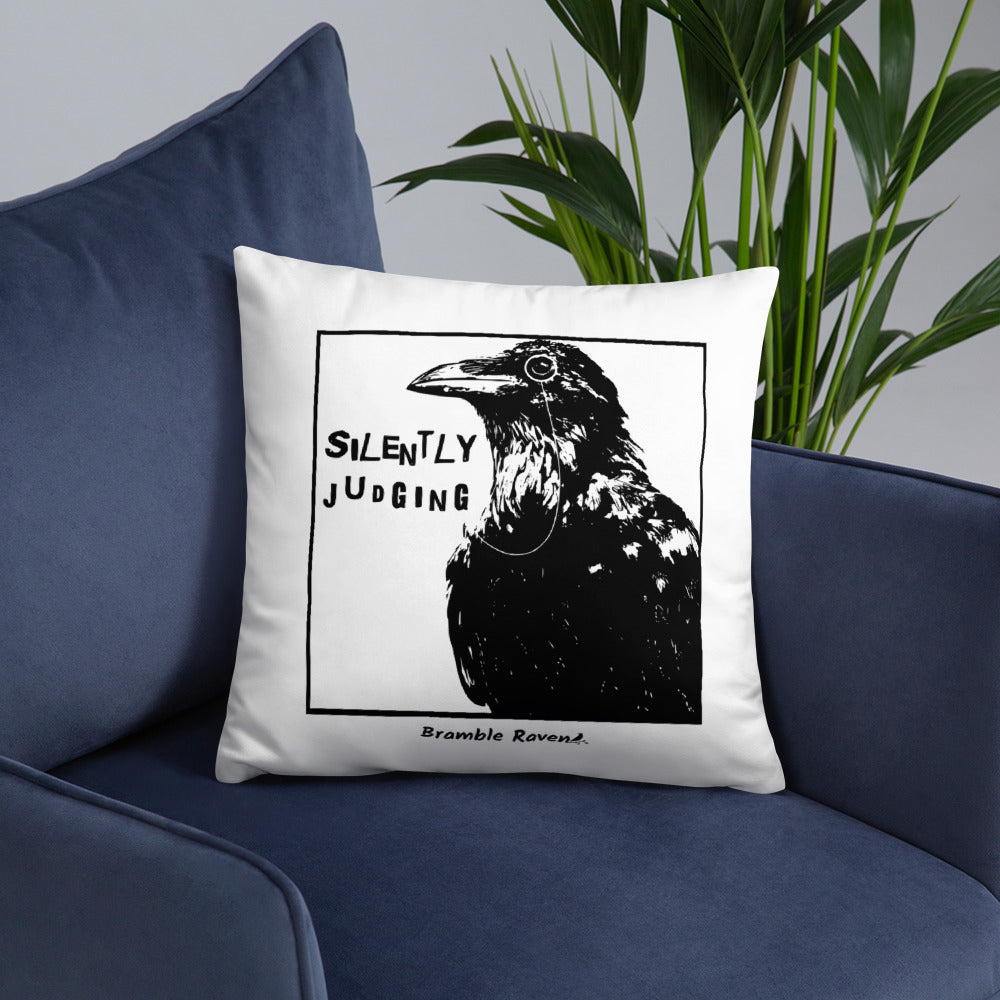 18 by 18 inch accent pillow with silently judging text by black crow wearing a monocle in a square on a white background.  Design has blue paint splatters on reverse side of pillow. Shown on blue sofa.