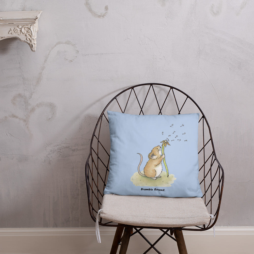 Original Dandelion Wish design of cute watercolor mouse blowing dandelion seeds on a light blue background. Double-sided image on 18 by 18 inch pillow. Shown on wicker chair.