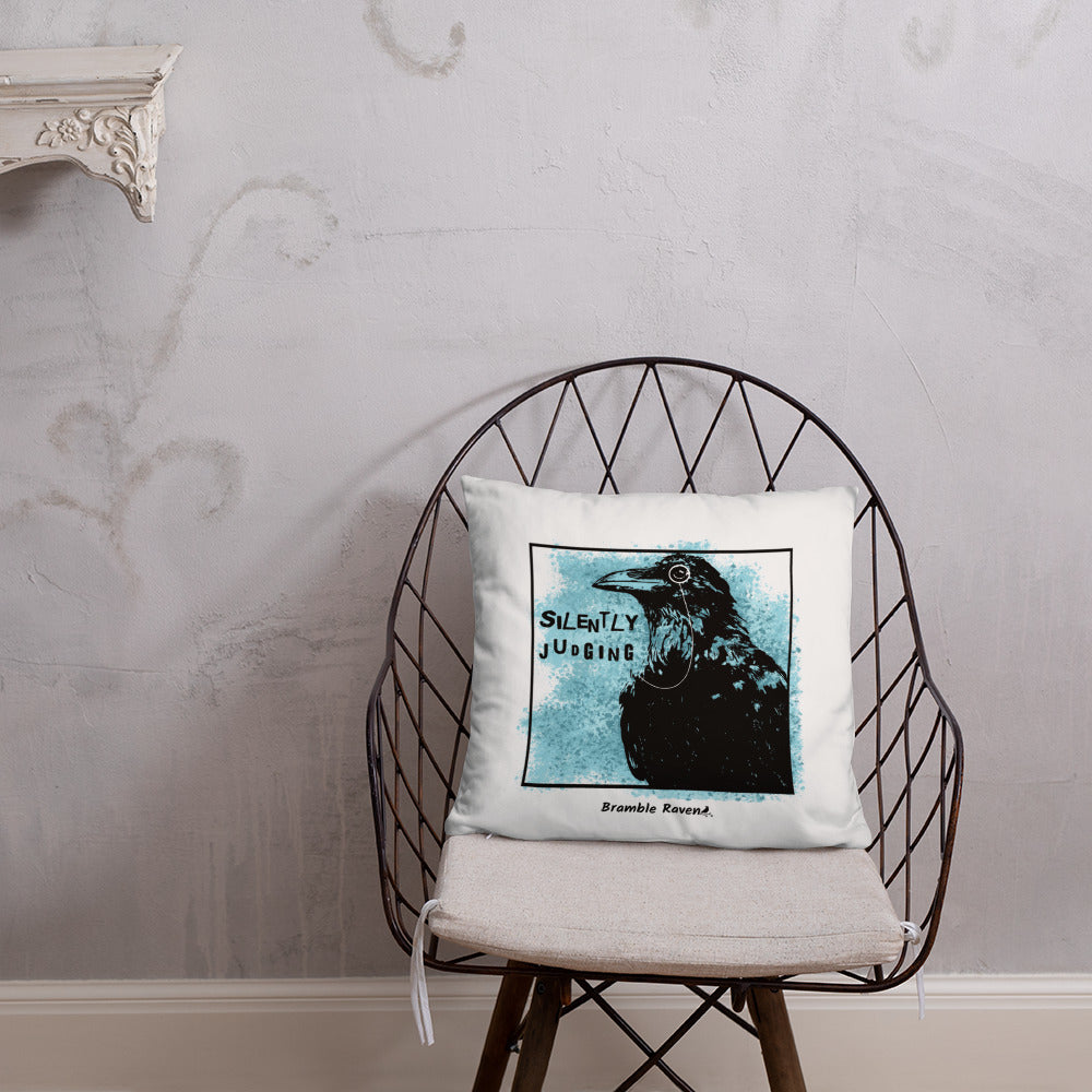 18 by 18 inch accent pillow with silently judging text by black crow wearing a monocle in a square with blue paint splatters on a white background.  Design on both sides of pillow. Shown on wicker chair.