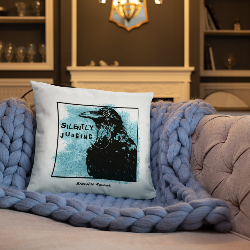 18 by 18 inch accent pillow with silently judging text by black crow wearing a monocle in a square with blue paint splatters on a white background.  Design on reverse side of pillow has no blue paint splatters. Shown on blue knitted blanket on tan sofa.
