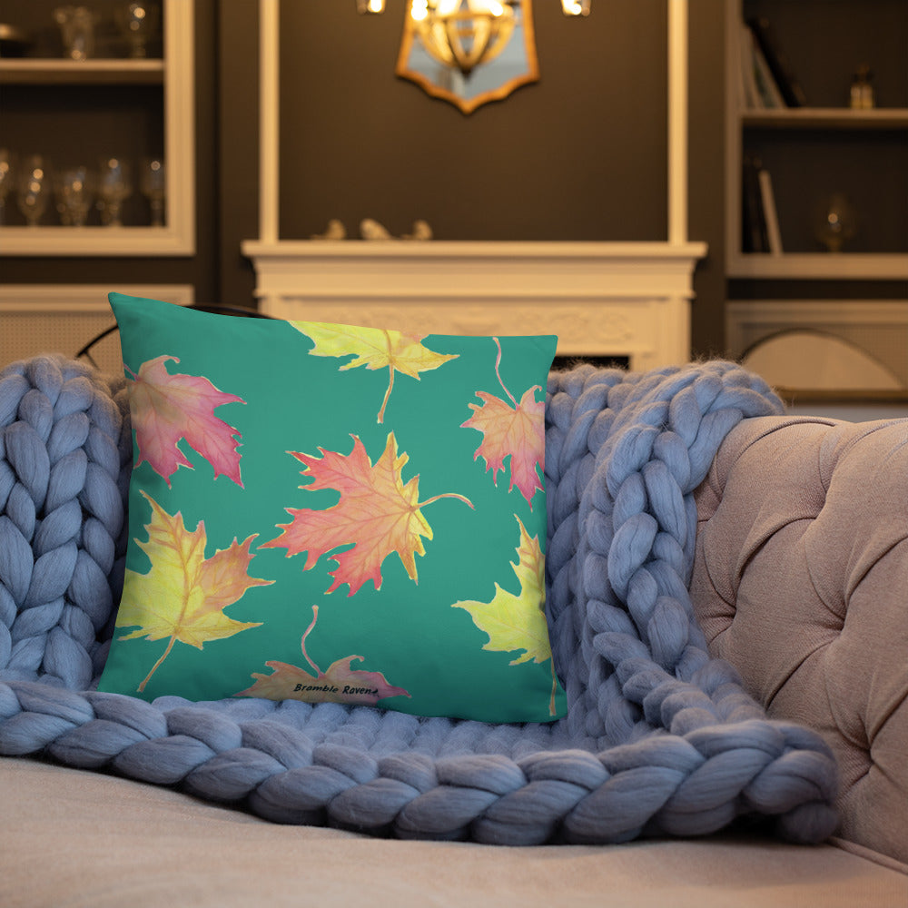 18 by 18 inch accent pillow featuring watercolor fall leaves on a dark green background. Pillow is double-sided. Larger patterned leaves on the back. Smaller patterned leaves on the front. Shown on a blue knitted blanket on a tan sofa.