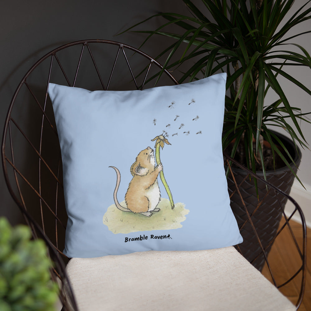 Original Dandelion Wish design of cute watercolor mouse blowing dandelion seeds on a light blue background. Double-sided image on 18 by 18 inch pillow. Shown on wicker chair with potted plant.